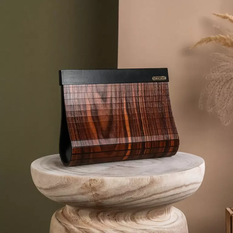 Charmant, flamboyant en vol zelfvertrouwen - dat is Chinira. Alle ogen zijn op haar gericht, waar ze ook heen gaat.

Deze clutch accentueert jouw outfits en is jouw beste vriendin tijdens lange avonden in de stad. Dankzij de natuurlijke, moderne uitstraling zijn alle ogen op jou gericht terwijl je jouw belangrijkste spullen altijd bij de hand hebt.

Materialen

Dankzij een geraffineerd ontwerp van smalle latten op het buigpunt heeft deze tas een complete laag van rozenhout. De houten onderdelen zorgen niet alleen voor de extravagante uitstraling, maar garanderen ook dat je een echt uniek en exclusief model draagt bij je draagt.

De binnenkant van de tas is bekleed met Italiaans nubuckleer. Dit voelt niet alleen zacht aan, maar is ook nog eens erg stevig.