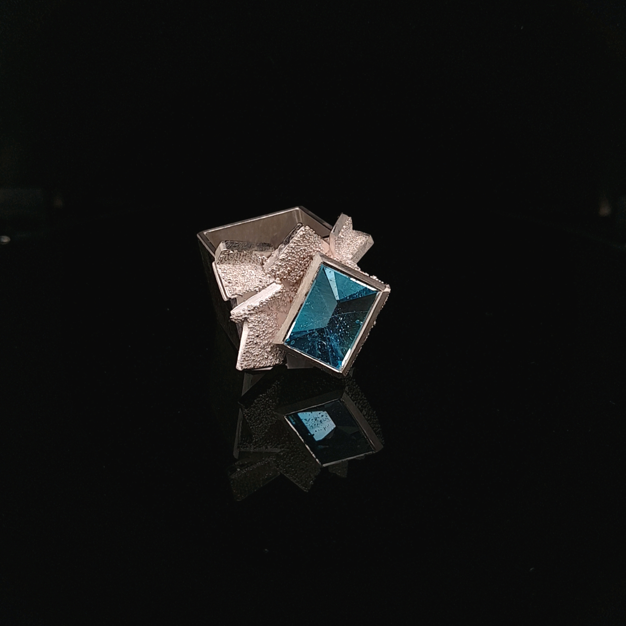 Ring gefertig in Silber 925, mit einem blauen Topas, im Spiegelschliff.
Die Ringschine ist Vierkant und granuliert.