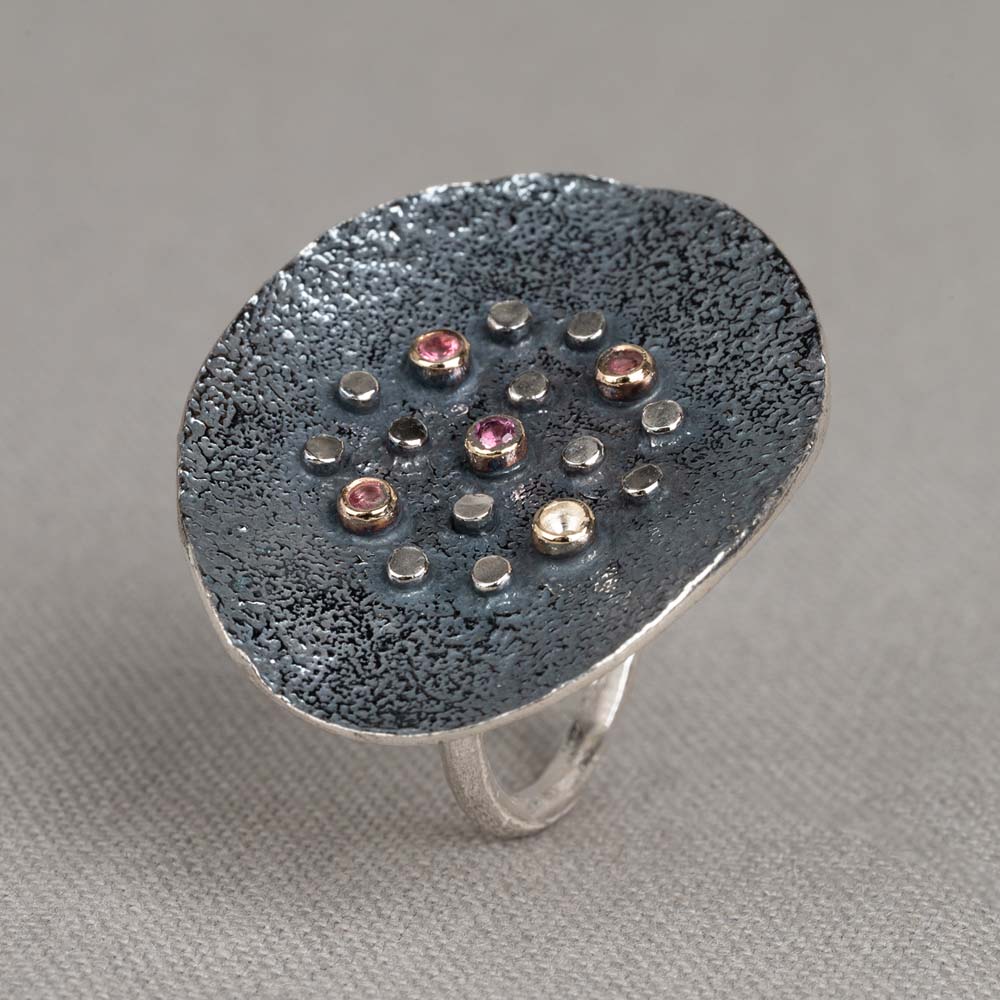 Ring gemaakt van 925 sterling zilver met daarop een schijf gemaakt van geoxideerd zilver. Hierop vind je kastjes gemaakt van 10 karaat en daarin de edelsteen Roze Toermalijn. Combineer deze ring met de bijpassende colliers en oorbellen.