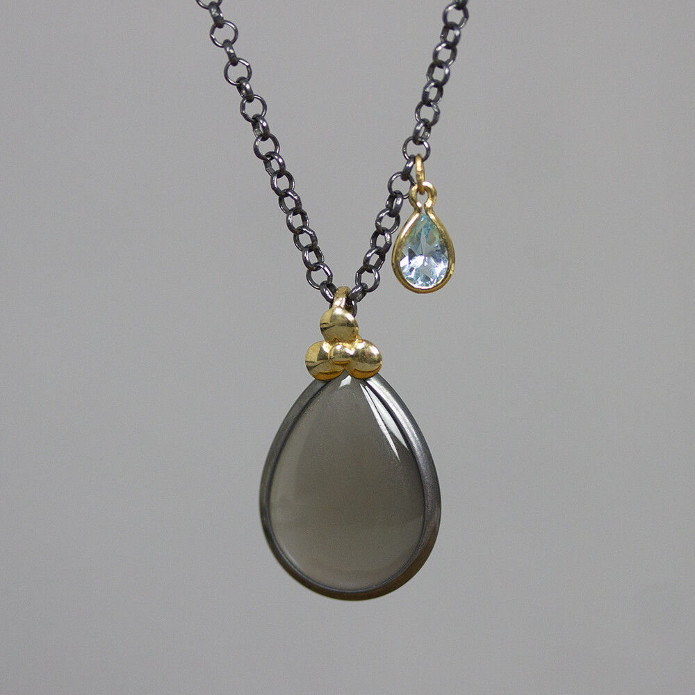 Collier gemaakt van 925 geoxideerd sterling zilver, met als grote edelsteen een grijze Maansteen en een kleine Blauwe Topaas. Boven de hanger zit een klavertje 3 van 14 karaat goud. Let op het model draagt dit collier in chocolade maansteen.