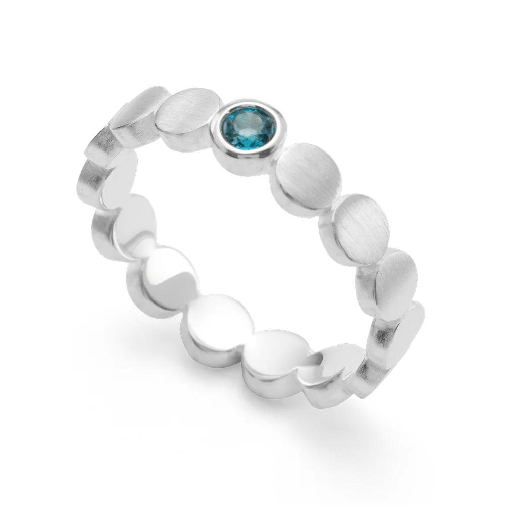 Deze moderne ring (ontwerp Nadine Resch) is samengesteld uit ronde, naast elkaar geplaatste decoratieve elementen, bekroond door een stralend blauwe topaas (0,14 karaat).