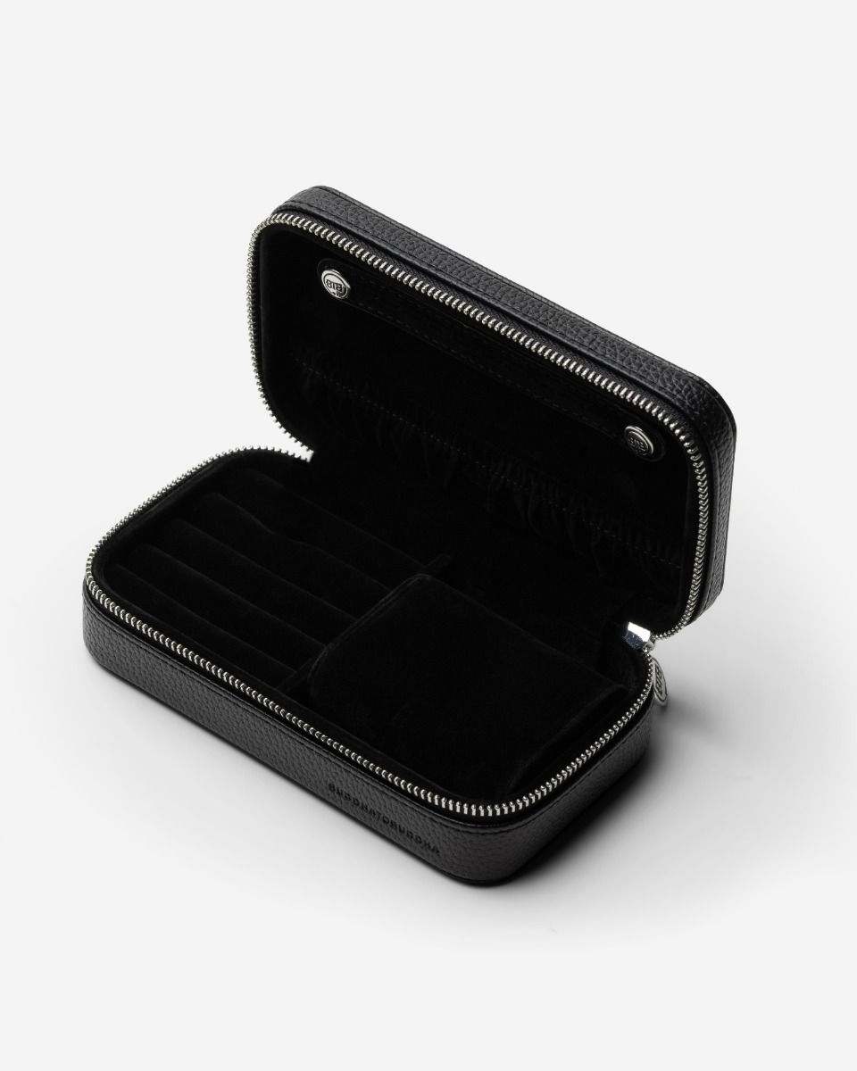 De Jewelry Travel Case Black Medium biedt een luxueuze en praktische manier om je sieraden overzichtelijk te bewaren, zowel thuis als onderweg. In de flap bevindt zich een afneembare strip met drukknopen voor het opbergen van 5 paar oorbellen, terwijl kettingen eromheen gehangen kunnen worden. Onderin biedt de case aan de linkerkant ruimte voor meerdere ringen, oorbellen en hangers, terwijl de rechterkant een uitneembare armband-rol bevat, ideaal voor meerdere armbanden. Voor grotere armbanden kan de armband-rol worden verwijderd om extra ruimte te creëren. De case is gemaakt van RCS-gecertificeerd gerecycled PU en de voering van GRS-gecertificeerd gerecycled polyester. De duurzame sieradenetui is afgewerkt met een zilverkleurige BTB-ritssluiting en BUDDHATOBUDDHA logo’s die in reliëf zijn aangebracht.