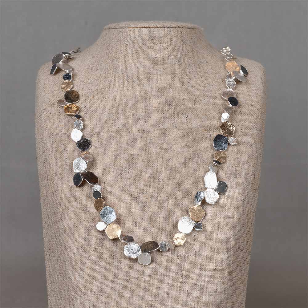 Dit collier valt op door de speelse compositie van kleine, organisch gevormde elementen. De schakels combineren 925 sterling zilver, geoxideerd zilver en verguld zilver. De licht gehamerde structuren geven elk element een ambachtelijke uitstraling en laten het licht subtiel weerkaatsen. Door de variatie in vormen, tinten en formaten ontstaat een levendig en elegant geheel dat soepel om de hals valt. Een opvallend maar verfijnd collier dat elke outfit een moderne en stijlvolle uitstraling geeft.