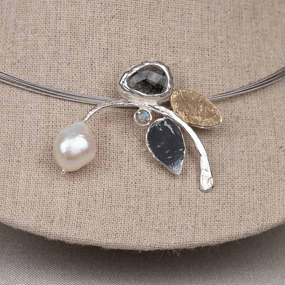 Deze hanger is gemaakt van 925 sterling zilver en combineert verschillende texturen, tinten en edelstenen in een speels organisch ontwerp. De hanger is verrijkt met een zachte glans van een Parel , de karaktervolle uitstraling van Black Rutiel en een schittering van Labradoriet . Elementen van geoxideerd zilver en verguld zilver zorgen voor een subtiel contrast tussen lichte, donkere en warme accenten. De licht gehamerde vormen geven het sieraad een ambachtelijke uitstraling en laten elke steen prachtig tot zijn recht komen.
