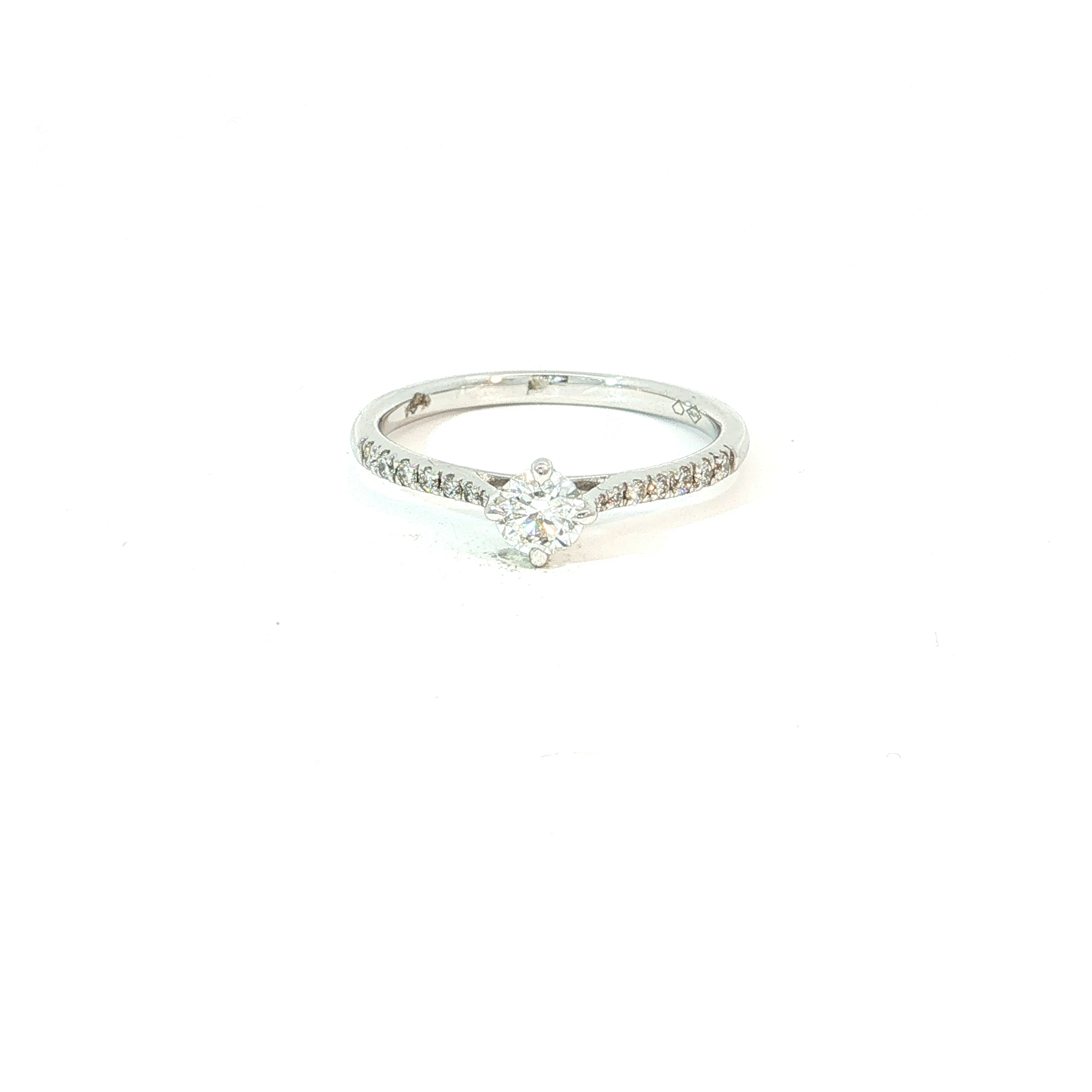Een elegante verlovingsring met een heldere, verfijnde uitstraling. De centrale briljant vormt het stralende middelpunt en wordt extra geaccentueerd door fijne briljanten die over de schouders van de ring zijn gezet.
Het 18 kt witgoud versterkt de frisse schittering van de diamanten en geeft het ontwerp een luxueuze, moderne uitstraling. De slanke ringband en de open zetting zorgen voor een licht en vrouwelijk karakter.
Een tijdloos juweel dat klasse en romantiek perfect combineert &mdash; ideaal als verlovingsring voor wie houdt van verfijnde luxe en maximale schittering.